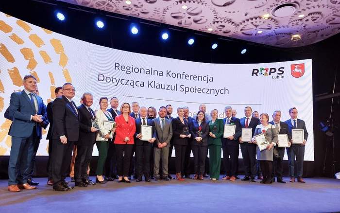 Pokaż zdjęcie: Na scenie na tle białego baneru z napisem Regionalna Konferencja Dotycząca Klauzul Społecznych pozuje duża grupa osób elegancko ubranych w średnim wieku. Część z nich trzyma w ręku dyplomy