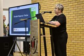 Pokaż zdjęcie: kobieta w okularach, która stoi przy mównicy i przemawia do mikrofonu. Na ekranie obok niej wyświetlana jest prezentacja z napisem „Ośrodek Wsparcia i Testów w Białymstoku”. Tło stanowi żółta, ceglana ściana. Na mównicy widoczny jest symbol przedstawiający postacie trzymające serce.