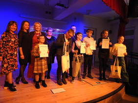 Pokaż zdjęcie: Na scenie stoi grupa szczęśliwych, uśmiechniętych młodych osób w rękach trzymają dyplomy i nagrody. Obok nich stoją osoby dorosłe.