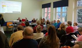 Pokaż zdjęcie: duża sala po środku siedzą osoby i oglądają slajdy które są wyświetlane na ścianie. Przy ścianie stoją stoliki przy których siedzą osoby i omawiają projekty PFRON 