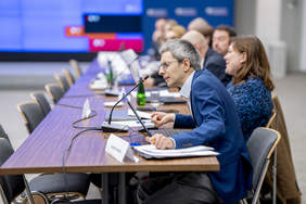 Pokaż zdjęcie: Mężczyzna w okularach i granatowej marynarce. Siedzi na krześle i mówi co mikrofonu, który stoi na stole przed nim. W tle inni siedzący przy stole ludzie.