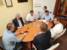 Pokaż zdjęcie: Przy owalnym stole siedzi sześciu mężczyzn. Są ubrani w koszule lub garnitury. Na stole leżą dokumenty, pieczątki, długopisy, telefony. 