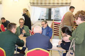 Pokaż zdjęcie: Przy stoliku siedzi 5 osób, dwie stoją przy stoliku. Przy stole trwają rozmowy i konsultacje