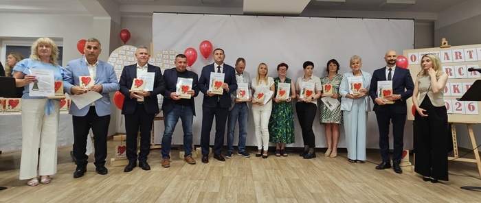 Pokaż zdjęcie: Grupa laureatów stoi na scenie z dyplomami i statuetkami z czerwonym sercem podczas gali Otwarte Serca 2026. W tle białe tło, czerwone balony oraz tablica z nazwą wydarzenia. Wszyscy pozują do pamiątkowego zdjęcia.  