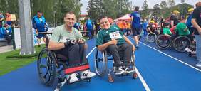 Pokaż zdjęcie: Dwóch panów siedzi na ręcznych wózkach inwalidzkich. Na koszulkach mają naklejki zawodników sportowych. W tle widać tłum uczestników olimpiady. 