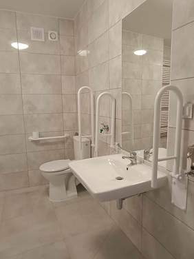 Pokaż zdjęcie: Zdjęcie fragmentu łazienki, zlew, lustro, toaleta wraz z urządzeniami umożliwiającymi osobie z niepełnosprawnością skorzystanie z toalety.