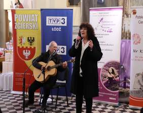 Pokaż zdjęcie: na zdjęciu występ artystyczny osoba siedzi na krześle i gra na gitarze obok stoi Pani trzyma mikrofon i śpiewa dla publiczności