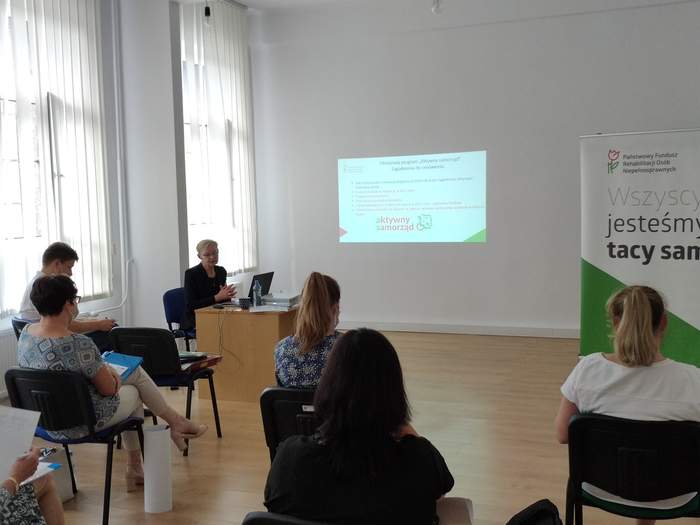 Pokaż zdjęcie: w pomieszczeniu przy biurku z laptopem siedzi kobieta ubrana na czarno w tle na ścianie wyświetlona jest prezentacja na sali znajduje się jeszcze kilka osób siedzących na krzesłach