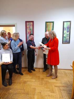 Pokaż zdjęcie: Kobieta w czerwonej sukience wręcza dyplom mężczyźnie, wokoło stoją inne osoby