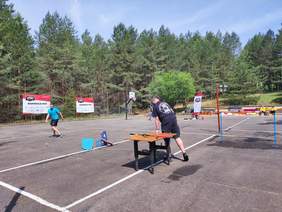 Pokaż zdjęcie: Dwóch mężczyzn stoi na boisku  boisku Jeden z nich stoi przy stole z ustawionym sprzętem, drugi idzie w stronę upuszczonej piłeczki. W tle widoczne są banery z napisem START „Wspierają nas”  popraw teks stylistycznie i gramatycznie 