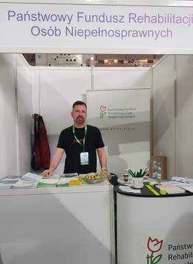 Pokaż zdjęcie: Mężczyzna stojący za stoiskiem Państwowego Funduszu Rehabilitacji Osób Niepełnosprawnych (PFRON) na targach. Na ladzie przed nim znajdują się broszury, ulotki, miska z cukierkami oraz gadżety reklamowe. W tle logo i nazwa PFRON na ściance wystawienniczej. Mężczyzna ubrany w czarną koszulkę i identyfikator na zielonej smyczy.