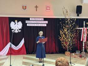 Pokaż zdjęcie: Kobieta w niebieskiej sukience przemawia do mikrofonu, trzymając czerwoną teczkę. W tle widoczne są godło Polski, biało-czerwona flaga, brzozowe dekoracje oraz krzyż z biało-czerwoną wstążką.