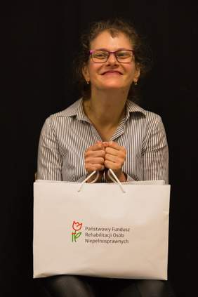 Pokaż zdjęcie: Uśmiechnięta kobieta w dłoniach trzyma białą torbę z napisem Państwowy Fundusz Rehabilitacji Osób Niepełnosprawnych.