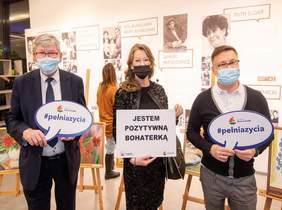 Pokaż zdjęcie: na zdjęciu: w tle prace malarskie na środku stoją trzy osoby które trzmają tabliczki z napisem pełnia życia oraz jestem pozytywną bohaterką 