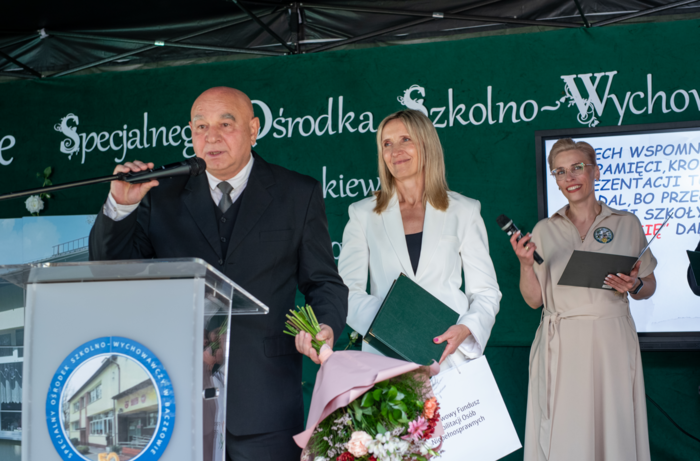 Pokaż zdjęcie: Na scenie przy mównicy stoi mężczyzna w garniturze, w ręku trzyma bukiet kwiatów, obok niego stoją dwie kobiety.