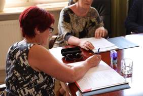 Pokaż zdjęcie: Przy stole siedzi kobieta w dłoni trzyma długopis podpisuje dokumenty.