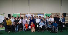 Pokaż zdjęcie: Kobiety i mężczyźni, w tym osoby z niepełnosprawnością, m.in. na wózkach inwalidzkich, pozują do zdjęcia w sali gimnastycznej