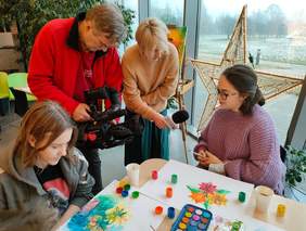 Pokaż zdjęcie: na zdjęciu dwie dziewczynki malują obrazy ustami obok dziennikarka telewizji TOYA  przeprowadza wywiad a kamerzysta kręci  materiał zdjęciowy