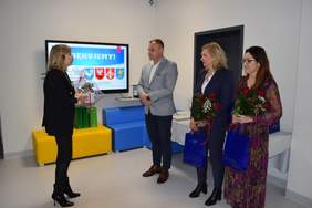 Pokaż zdjęcie: W sali w której na ścianie wisi duży telewizor, pod ścianą stoja siedziska w kolorze żółtym i niebieskim, stoją cztwery osoby, po lewej kobieta w garniturze, skierowana twarza do trójki osób: mężczyzny i dwóch kobiet, kobiety trzymają w ręku prezenty i granatowe torby reklamowe.