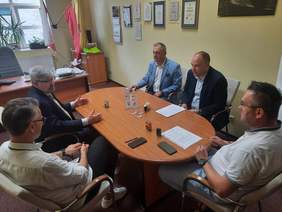 Pokaż zdjęcie: Przy owalnym stole siedzi pięciu mężczyzn, którzy ze sobą rozmawiają. Na stole leżą dokumenty, pieczątki, woda mineralna. 