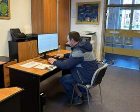 Pokaż zdjęcie: Za biurkiem siedzi mężczyzna przed komputerem patrzy w monitor. W pokoju jest szara wykładzina na ścianach wiszą kolorowe obrazy.