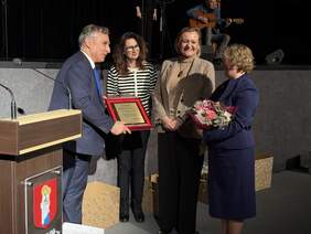 Pokaż zdjęcie: Cztery osoby stoją na scenie podczas uroczystości. Mężczyzna w garniturze wręcza czerwone etui z dyplomem kobiecie trzymającej bukiet kwiatów. Obok stoją dwie kobiety obserwujące moment wręczenia nagrody. Z lewej strony pulpit z mikrofonem.