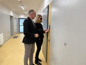 Pokaż zdjęcie: Mężczyzna w czarnej marynarce szarych spodniach i w okularach, trzyma za rękę  kobietę wskazując jej elementy na ścianie. Kobieta jest blondynką, nosi ciemne okulary przeciwsłoneczne jest ubrana w czarną bluzkę, czarne obcisłe spodnie i czarne