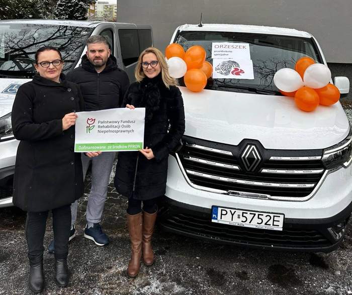Pokaż zdjęcie: Dwie kobiety trzymają w dłoniach naklejkę z logo PFRON informującym o dofinansowaniu ze środków PFRON. Z tyłu stoi mężczyzna. W tle dwa busy. Jeden z nich udekorowany balonami. Na szybie pojazdu naklejka z logo Przedszkola Specjalnego „Orzeszek”.