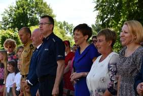 Pokaż zdjęcie: Na tle drzew liściastych grupa kilkunastu uśmiechniętych osób kieruje swój wzrok w jedną stronie. W tej grupie znajdują się między innymi: dzieci, przedstawiciele służb mundurowych oraz różnych innych instytucji.