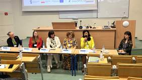 Pokaż zdjęcie: Przy stole w auli siedzi sześć kobiet: moderatkorka i panelistki. Kobiety wypowiadają się do mikrofonu.