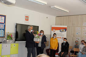 Pokaż zdjęcie: Dwie osoby stoją przy czarnej tablicy, mężczyźnie wręczane są kwiaty