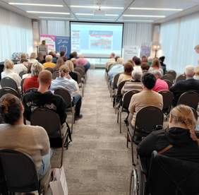 Pokaż zdjęcie: Zdjęcie przedstawia widok z tyłu na salę konferencyjną wypełnioną uczestnikami siedzącymi na krzesłach i skierowanymi w stronę ekranu projekcyjnego. Na ekranie widać prezentację z logo J-elita, Polskiego Towarzystwa Wspierania Osób z Nieswoistymi Zapaleniami Jelita. Z tyłu sali stoi kobieta, prawdopodobnie prowadząca spotkanie. W pierwszym rzędzie widać osobę na wózku inwalidzkim.