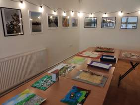 Pokaż zdjęcie: fragment pomieszczenia, na białych ścianach wiszą w rzędzie oświetlone fotografie na stołach są rozłożone prace plastyczne