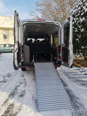 Pokaż zdjęcie: Na parkingu stoi ustawiony tyłem bus z otwartymi drzwiami i rozłożoną platformą wspomagającą transport osób na wózkach inwalidzkich.