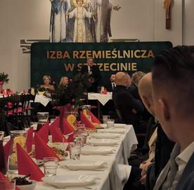 Pokaż zdjęcie: Uroczysta sala podczas Spotkania Opłatkowo-Noworocznego. Na pierwszym planie udekorowane stoły z czerwonymi serwetkami i świątecznymi ozdobami, w tle widoczny mówiący prelegent, a za nim mozaika Świętej Rodziny.