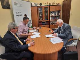 Pokaż zdjęcie: Przy owalnym stole siedzi trzech mężczyzn. Podpisują różne dokumenty. Na stole leżą umowy, pieczątki, długopisy, telefony. 