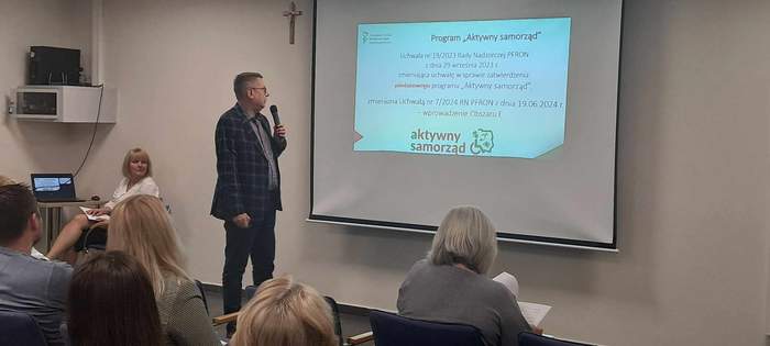 Po lewej stronie stoi mężczyzna i trzyma mikrofon w dłoni. Przemawia, patrząc na ekran, który wisi na ścianie i na slajd wyswietlony na nim. Slajd dotyczy programu PFRON "Aktywny samorząd"