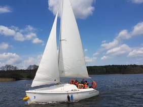 Pokaż zdjęcie: Na jeziorze płynie łódka z dwoma żaglami. W łódce siedzi 5 osób w kapokach. W tle lasy i błękitne troszkę zachmurzone niebo.