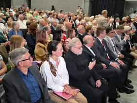 Pokaż zdjęcie: Licznie zgromadzona publiczność w różnym wieku wypełnia salę podczas wydarzenia. W pierwszym rzędzie siedzą elegancko ubrani uczestnicy, w tym mężczyźni w garniturach i kobieta w białej bluzce. Wszyscy siedzą blisko siebie i patrzą w lewą stronę, skupiając uwagę na scenie.