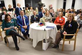 Pokaż zdjęcie: Zdjęcie sali z dużą ilością okrągłych stołów przy których siedzą kobiety i mężczyźni. Zbliżenie na stół przy którym siedzi 6 osób, pomiędzy stolikami stoi mężczyzna z kamerą