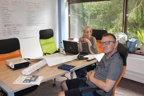 Pokaż zdjęcie: Kobieta i mężczyzna siedzą przy biurku. Na nim  stoi laptop. Za nimi tablica z zapisanymi informacjami