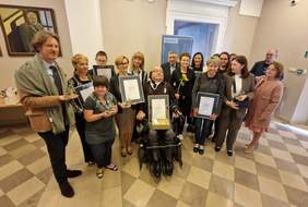 Pokaż zdjęcie: Grupa uśmiechniętych osób. W rękach trzymają kwiaty, dyplomy i statuetki. Wśród osób na zdjęciu są osoby z różnymi niepełnosprawnościami, m.in. na wózku oraz osoby z Zespołem Downa.