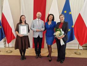 Pokaż zdjęcie: Cztery osoby stoją na tle flag Polski i Unii Europejskiej. Kobieta po lewej trzyma certyfikat, mężczyzna obok niej statuetkę, a kobieta po prawej bukiet kwiatów. Wszyscy uśmiechają się do zdjęcia.