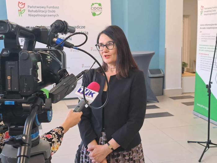 Wiceprezes Zarządu PFRON - Dorota Habich stoi przed kamerą i udziela wywaidu. W tle ścianka PFRON.