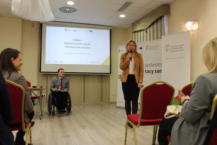 Pokaż zdjęcie: Zdjęcie z sali konferencyjnej, na pierwszym planie kobieta w beżowej marynarce i czarnej spódnicy, stoi na środku sali i przemawia do publiczności. W tle mężczyzna na wózku prowadzący konferencję, na ścianie ekran z prezentacją