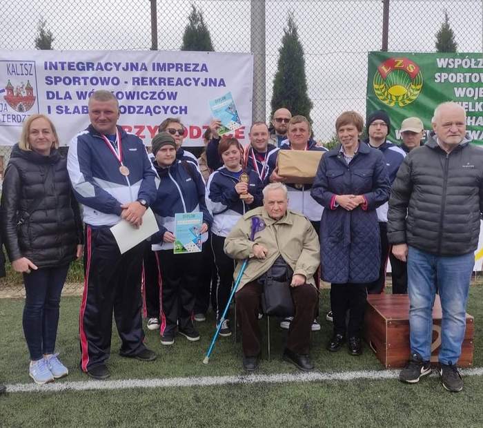 Pokaż zdjęcie: Na boisku widoczni są ustawieni w rzędzie zawodnicy rywalizujący w ramach XVIII Wielkopolskiej Imprezy Sportowo-Integracyjnej Osób Niepełnosprawnych. Są to zarówno kobiety, jak i mężczyźni. Część z nich ma na sobie medale. Wśród grupy zawodników znajdują się również inne osoby ubrane w kurtkach. W tle możemy dostrzec także banery informujące o nazwie i organizatorze wydarzenia oraz o źródłach jego finansowania.