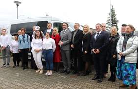 Pokaż zdjęcie: Grupa kilkunastu osób stoi na placu wyłożonym kostką brukową, obok białego busa, dostosowanego do przewozu osób z niepełnosprawnością. W tle widoczne są lampy uliczne oraz jedno wysokie drzewo iglaste.