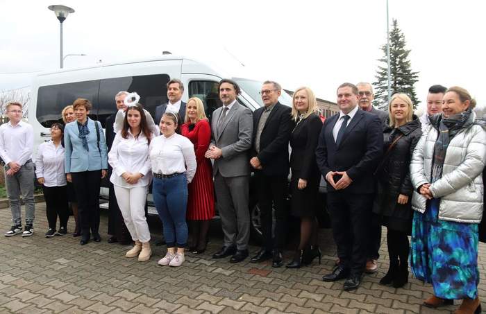 Pokaż zdjęcie: Grupa kilkunastu osób stoi na placu wyłożonym kostką brukową, obok białego busa, dostosowanego do przewozu osób z niepełnosprawnością. W tle widoczne są lampy uliczne oraz jedno wysokie drzewo iglaste.