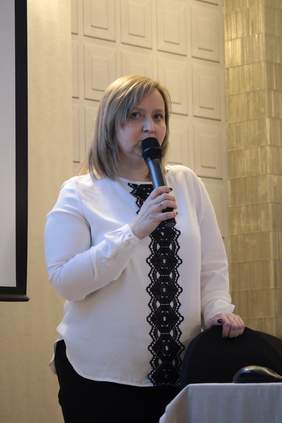 Pokaż zdjęcie: Przy stole konferencyjnym stoi kobieta ze średniej długości włosami, w białej bluzce z czarną aplikacją przez środek bluzki, w prawej ręce trzyma mikrofon