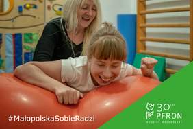 Pokaż zdjęcie: Podopieczna Fundacji w trakcie zajęć. Na zdjęciu ćwicząca uśmiechnięta  dziewczynka. Obok niej terapeuta